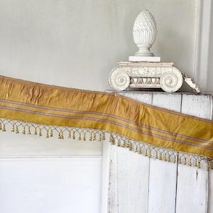 Puede incluir: Un borde de tela largo de color amarillo mostaza con rayas horizontales en tonos gris y blanco. El borde inferior está acabado con un fleco decorativo que consiste en pequeñas cuentas amarillas colgantes unidas a una delicada red tejida.