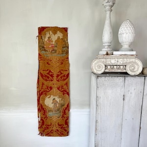 May include: A vintage tapestry with a red and gold background and a scene of two figures in a landscape. The tapestry is hanging on a white wall next to a white cabinet with two white decorative finials on top.