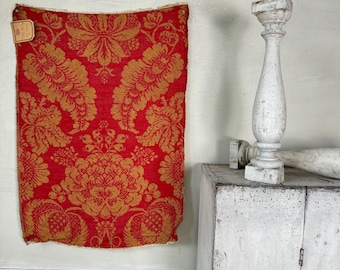 May include: A piece of red fabric with a gold floral pattern hangs on a white wall. The fabric has a tag attached to it. A white, weathered wooden pillar sits on a white cabinet.