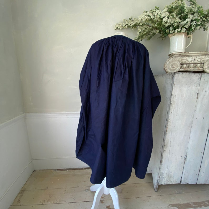 May include: A navy blue, loose-fitting, long tunic-style top with a gathered neckline. The top is made of a soft, drapey fabric and is hanging on a white stand.