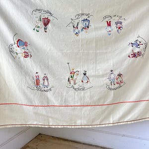 May include: A white linen tablecloth with red embroidered borders and colorful embroidered figures representing different regions of France. The figures are dressed in traditional clothing and each region is labeled with its name in French. The regions include: Lorraine, Provence, Champagne, Bretagne, Touraine, and Aquitaine.