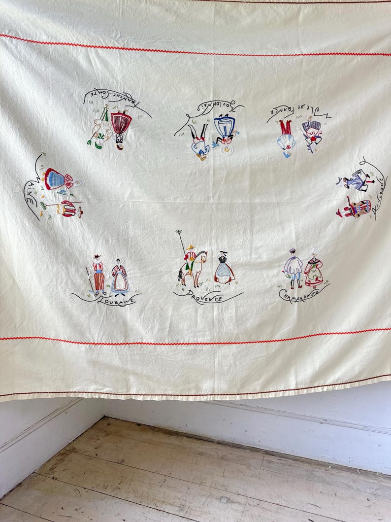 May include: A white linen tablecloth with red embroidered borders and colorful embroidered figures representing different regions of France. The figures are dressed in traditional clothing and each region is labeled with its name in French. The regions include: Lorraine, Provence, Champagne, Bretagne, Touraine, and Aquitaine.