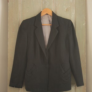 May include: A black blazer with a single button closure and two decorative pockets on the front. The blazer is hanging on a wooden hanger against a light green wooden door.