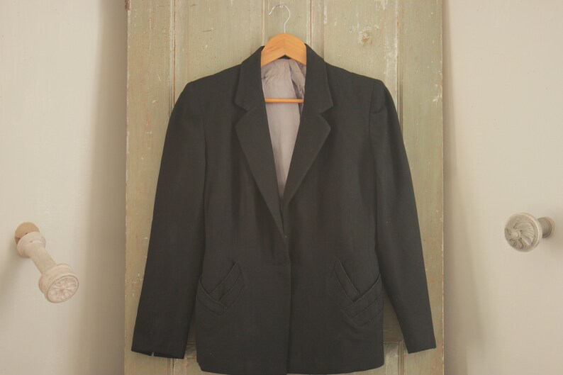 May include: A black blazer with a single button closure and two decorative pockets on the front. The blazer is hanging on a wooden hanger against a light green wooden door.