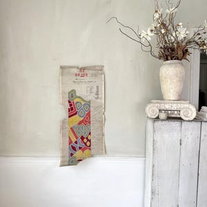 Puede incluir: Un documento vintage rasgado con patrones geométricos coloridos en rojo, amarillo, azul y verde cuelga de una pared verde claro. Un jarrón blanco con flores secas se encuentra encima de un armario blanco. El documento tiene el texto "ST. 26192".