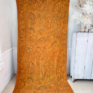 May include: An orange textile with a detailed floral pattern in shades of brown and gold. The fabric is draped, showcasing the intricate design of flowers, leaves, and other botanical elements. The textile is likely a decorative piece, such as a wall hanging or a throw.