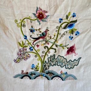 May include: Embroidered floral design on a white linen fabric. The design features a stylized tree with green leaves, pink and blue flowers, and a small bird with red, green, and blue feathers.