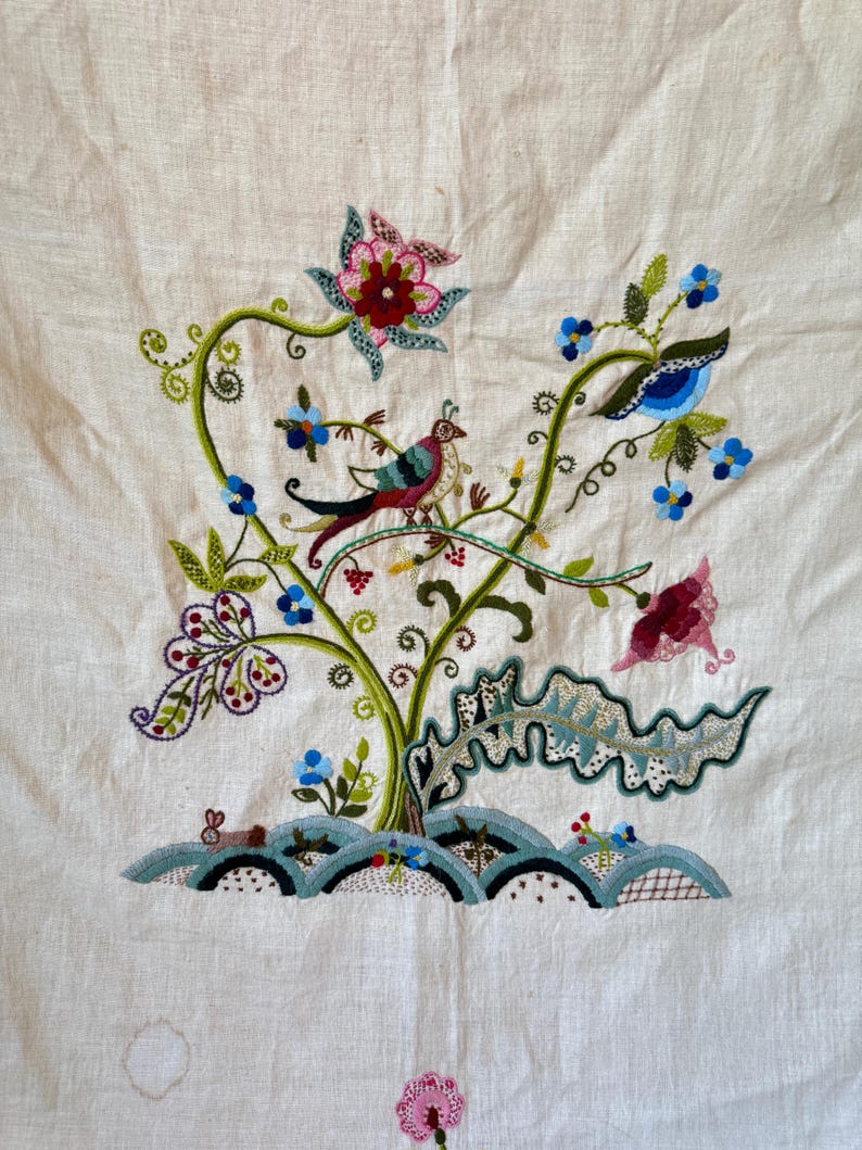 May include: Embroidered floral design on a white linen fabric. The design features a stylized tree with green leaves, pink and blue flowers, and a small bird with red, green, and blue feathers.