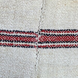May include: Close-up of a woven fabric with a red, black, and white striped pattern. The fabric is made of natural fibers and has a rough texture.