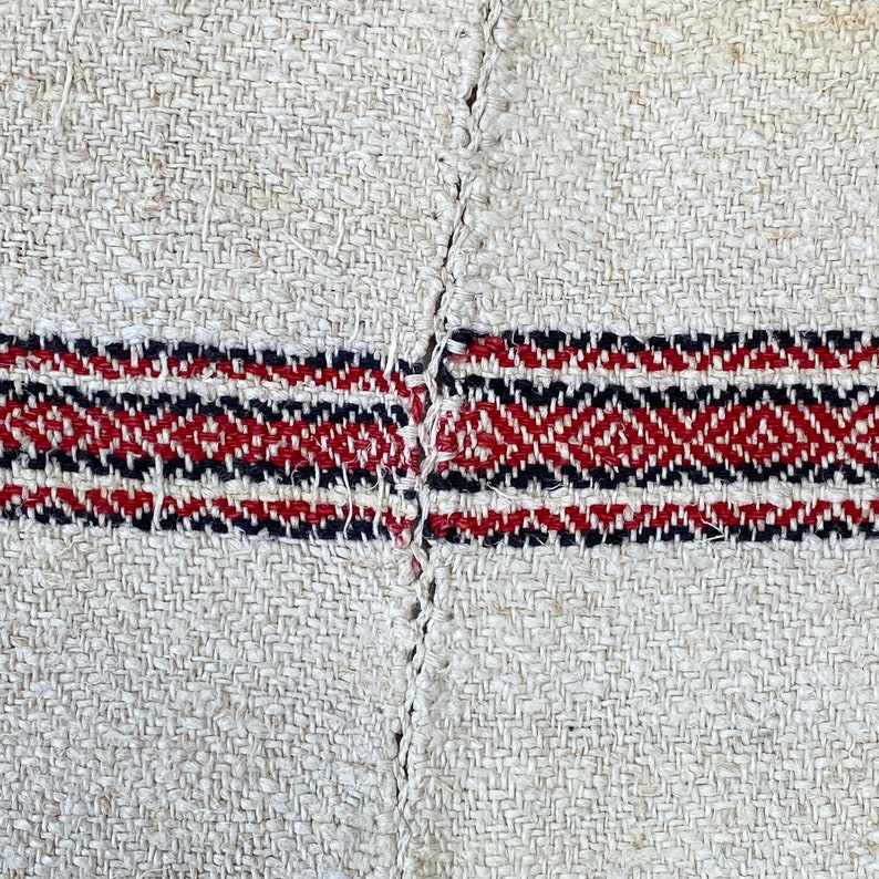 May include: Close-up of a woven fabric with a red, black, and white striped pattern. The fabric is made of natural fibers and has a rough texture.