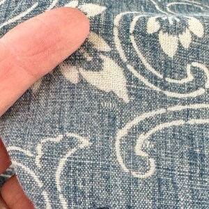 May include: Close-up of a blue and white fabric with a floral pattern. The fabric is textured and has a slightly faded look. The fabric is being held by a finger.