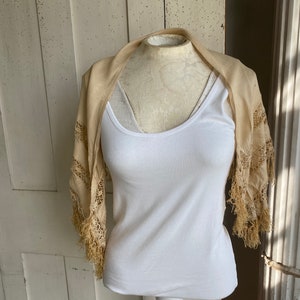 May include: A cream-colored lace shawl with fringe trim draped over a white tank top on a mannequin.