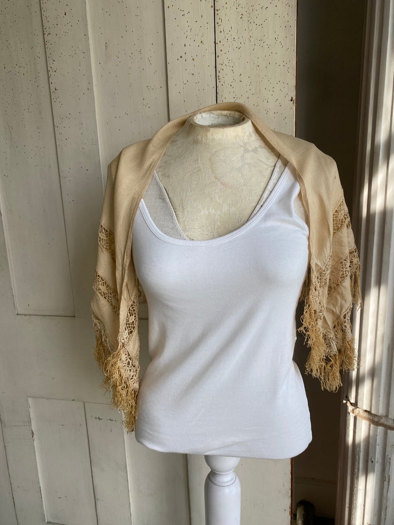 May include: A cream-colored lace shawl with fringe trim draped over a white tank top on a mannequin.