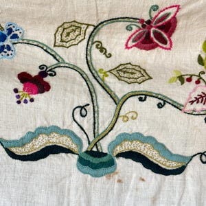 May include: A close-up of a white linen fabric with embroidered floral designs in shades of green, blue, pink, and red. The embroidery features a butterfly, leaves, and flowers.