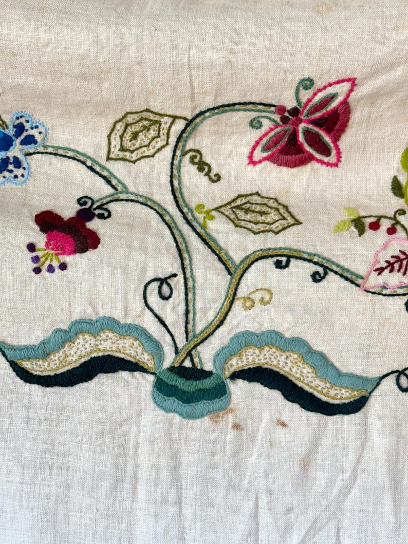 May include: A close-up of a white linen fabric with embroidered floral designs in shades of green, blue, pink, and red. The embroidery features a butterfly, leaves, and flowers.