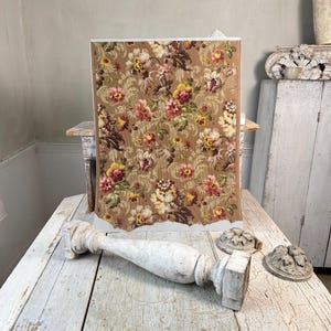 May include: A rectangular piece of vintage floral wallpaper with a brown background and colorful flowers. A distressed, white, wooden decorative element is in the foreground, along with two small, decorative, carved pieces.