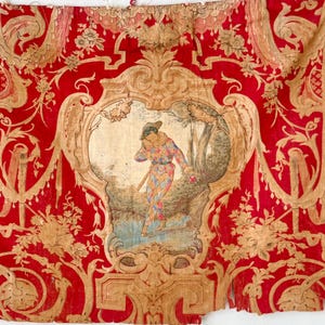 Könnte beinhalten: Ein Stück antiken Stoffes mit einem leuchtend roten Hintergrund und aufwendigen goldenen Blumenmustern. Das zentrale Design zeigt eine gerahmte pastorale Szene mit einer Figur in Harlekin-Kostüm. Der Stoff weist Alters- und Abnutzungserscheinungen auf.