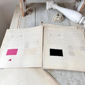 May include: Vintage textile design documents with handwritten notes and color swatches. The documents are on aged paper, with a pink and black swatch visible. The word "TISSAGE" is printed on one of the sheets. A distressed wooden table and decorative elements are in the background.
