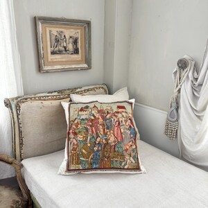 May include: A decorative pillow with a tapestry design featuring a medieval scene with figures in colorful clothing. The pillow is placed on a white bed with a light-colored headboard. A framed print hangs on the wall above the bed.