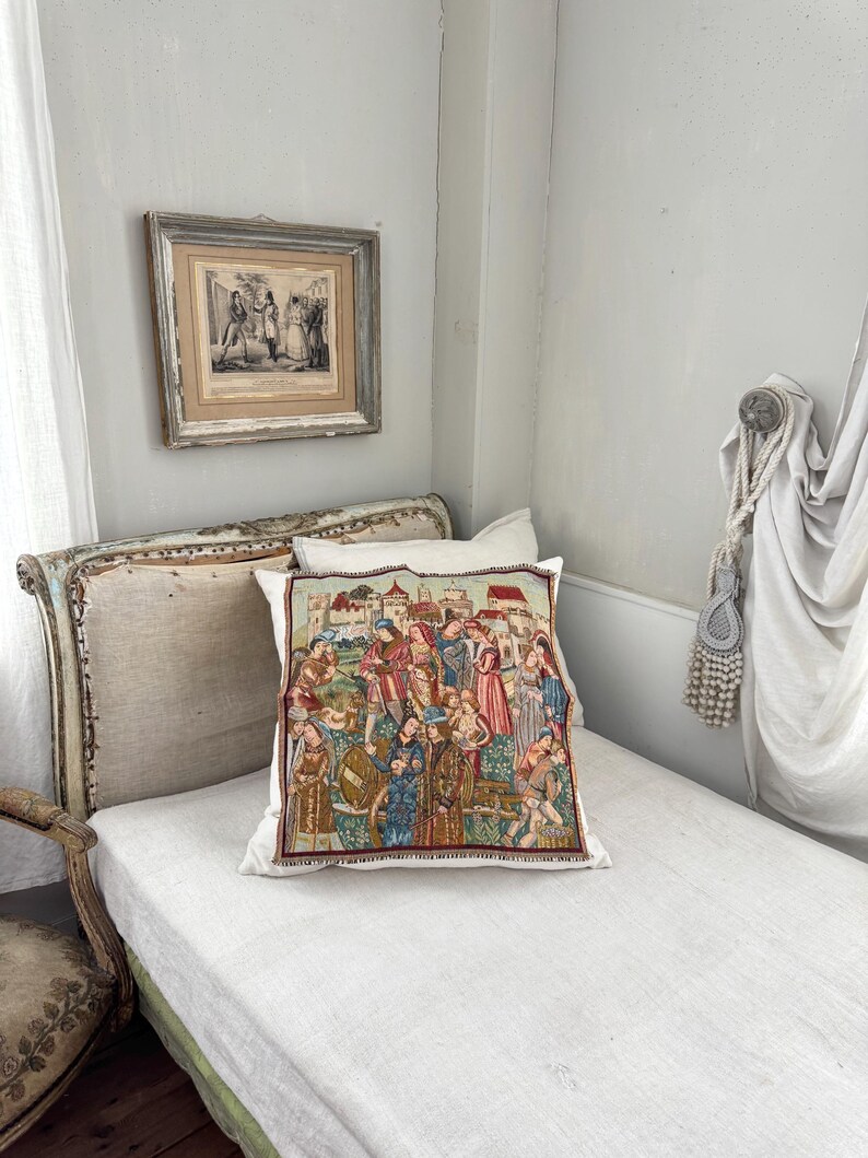 May include: A decorative pillow with a tapestry design featuring a medieval scene with figures in colorful clothing. The pillow is placed on a white bed with a light-colored headboard. A framed print hangs on the wall above the bed.