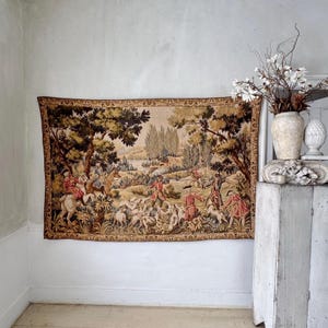 May include: A large tapestry depicting a hunting scene with figures on horseback, dogs, and people in period clothing. The tapestry features a landscape with trees, a lake, and a detailed border. The color palette includes beige, brown, green, and red.
