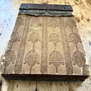 May include: A vintage wallpaper sample book with a brown embossed pattern of stylised leaves and a metal binding. The book is worn and has some damage.