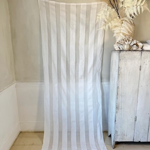 Puede incluir: Panel de cortina blanca transparente con rayas verticales. La cortina está colgada en una pared en una habitación con suelo de madera pintado de blanco y un armario pintado de blanco.