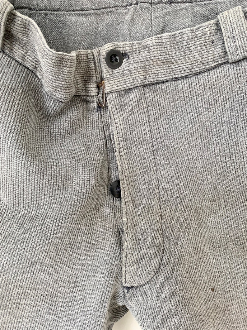 May include: Close-up of the front of a pair of gray pants with a button fly and a single button closure. The pants have a textured, ribbed fabric.