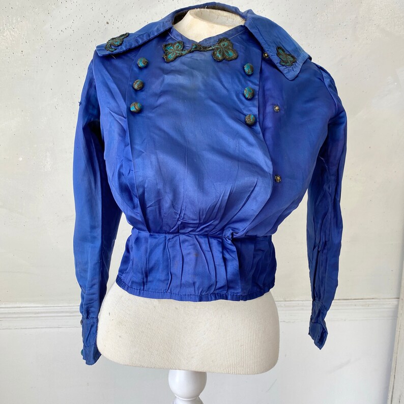 May include: A vintage cobalt blue blouse with long sleeves and a double-breasted design. The blouse features decorative buttons and ornate embellishments on the collar and neckline. The waist is cinched with pleats.