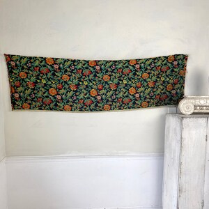 May include: A black fabric tapestry with a floral pattern in shades of orange, red, yellow, and green. The tapestry is hanging on a white wall.