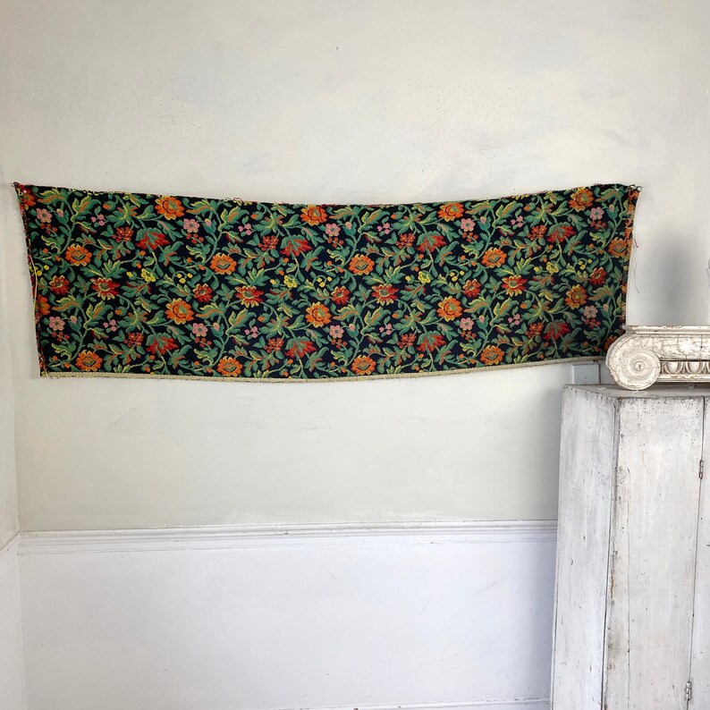 May include: A black fabric tapestry with a floral pattern in shades of orange, red, yellow, and green. The tapestry is hanging on a white wall.
