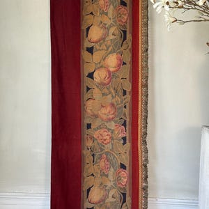 Peut inclure: Un long panneau de tapisserie avec un fond rouge et une bande verticale de tissu tissé présentant un motif de fruits, de fleurs et de feuilles dans des tons de brun, d'or et de vert. Le panneau a une bordure frangée.