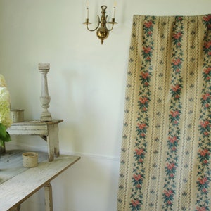 K&ouml;nnte beinhalten: Ein Vintage-Vorhang mit floralem Muster auf beigefarbenem Grund, roten und gr&uuml;nen floralen Akzenten und wei&szlig;em Saum. Der Vorhang h&auml;ngt in einem Raum mit wei&szlig;en W&auml;nden und einem Holztisch mit einer Vase mit wei&szlig;en Blumen.