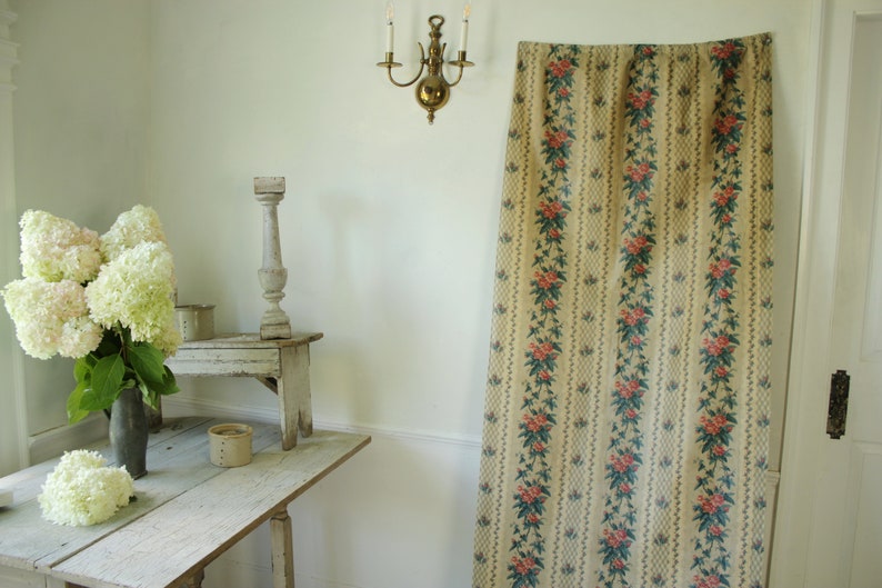K&ouml;nnte beinhalten: Ein Vintage-Vorhang mit floralem Muster auf beigefarbenem Grund, roten und gr&uuml;nen floralen Akzenten und wei&szlig;em Saum. Der Vorhang h&auml;ngt in einem Raum mit wei&szlig;en W&auml;nden und einem Holztisch mit einer Vase mit wei&szlig;en Blumen.