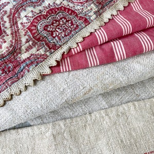 Op de afbeelding: Een stapel stof met een rood-wit gestreepte stof, een beige linnen stof en een rood-blauwe paisley stof met een witte gehaakte rand.