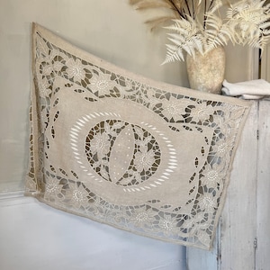 May include: A beige, openwork textile with a floral design. The textile is draped over a white cabinet. The floral pattern is centered with an oval shape and surrounded by a border of flowers and cut-out details. A vase with dried plants is in the background.