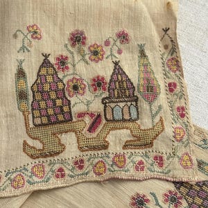 May include: Close-up of a beige textile with intricate cross-stitch embroidery. The design features stylized buildings, floral motifs in pink, yellow, and green, and a decorative border. The embroidery is detailed and colorful.