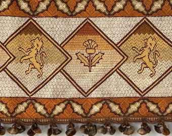 May include: A decorative textile with a repeating diamond pattern. The diamonds feature a lion and a thistle design in shades of gold, brown, and cream. The textile is trimmed with a patterned border and tassels.