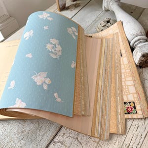 May include: An open vintage wallpaper sample book. The pages display various patterns, including a light blue page with white floral designs. Other pages show stripes and floral patterns in beige and pink tones. The book is on a distressed white wooden surface.
