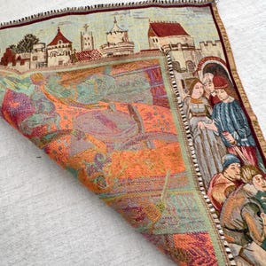 May include: A tapestry with a medieval scene featuring a townscape and figures in period clothing. The tapestry has a detailed border and is made of woven fabric. The color palette includes shades of orange, green, and blue.