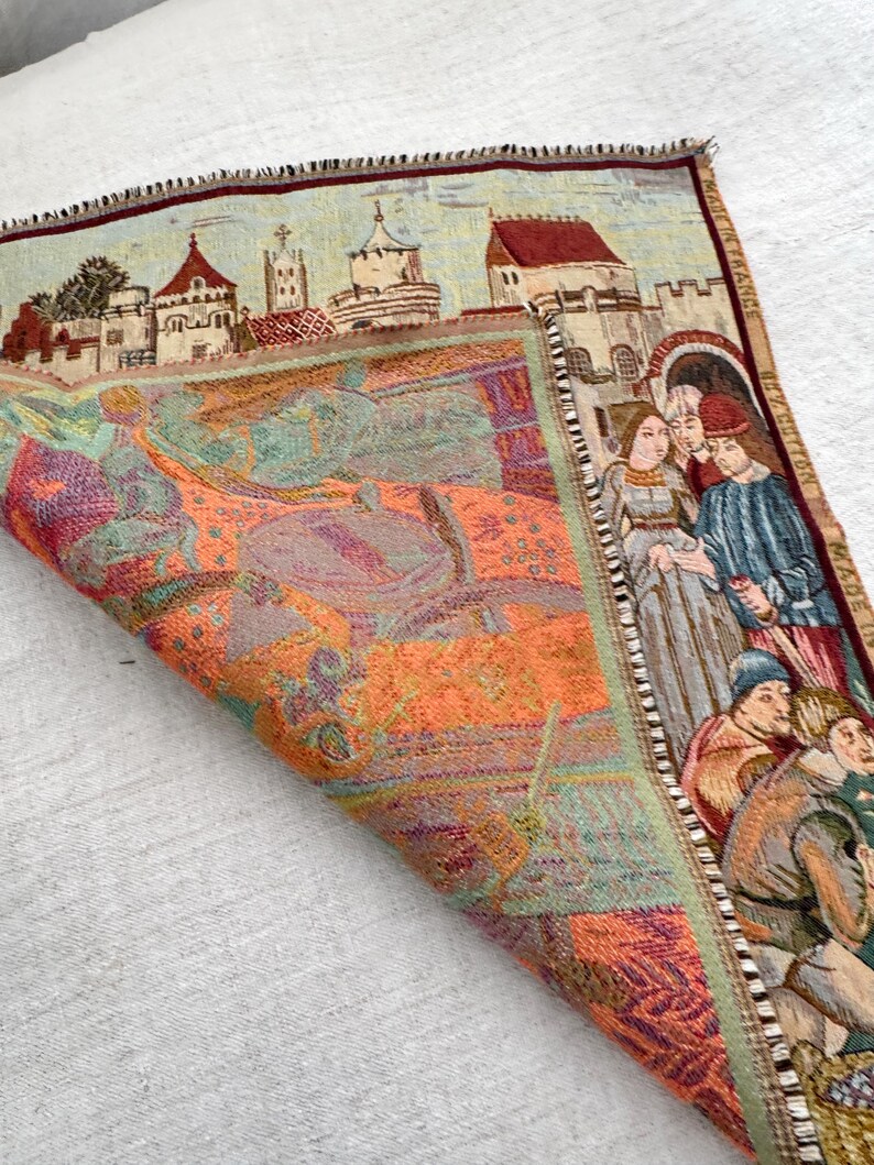 May include: A tapestry with a medieval scene featuring a townscape and figures in period clothing. The tapestry has a detailed border and is made of woven fabric. The color palette includes shades of orange, green, and blue.