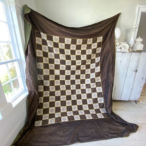 May include: A brown and white checkered fabric with a decorative pattern of sunbursts. The fabric has a fringed edge and is made of a soft, luxurious material.