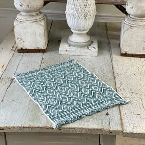 May include: A green and white woven placemat with a geometric pattern and frayed edges. The placemat is on a whitewashed wooden table.