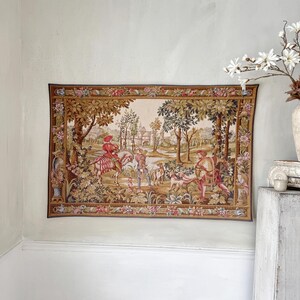 May include: A rectangular tapestry depicting a pastoral scene with figures, trees, and a building. The tapestry has a brown border with floral designs and is mounted on a light-colored wall. The scene includes a person on horseback and people with dogs.