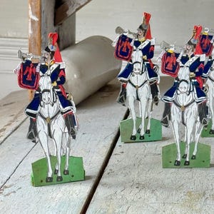 May include: A collection of vintage toy soldiers on horseback. The soldiers wear red, blue, and white uniforms and are mounted on white horses. Each figure holds a trumpet and a shield with a crest. The bases are green.