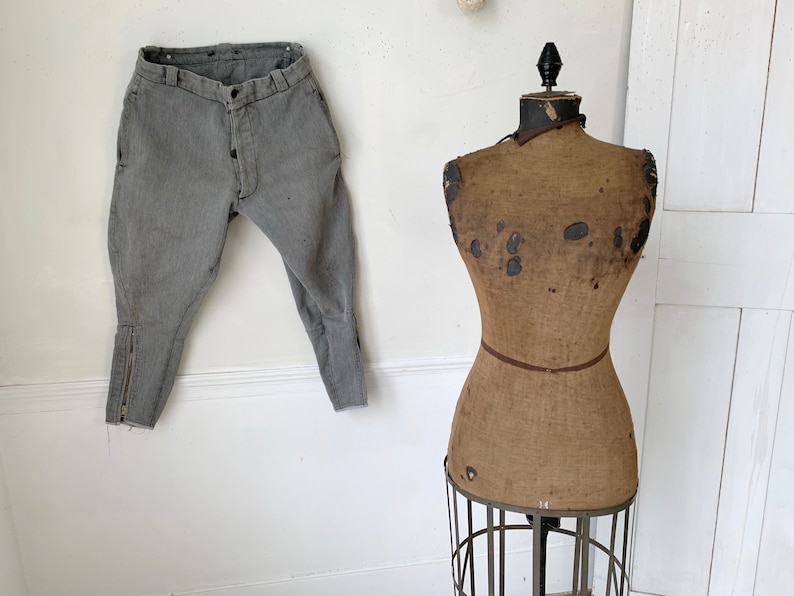 May include: A pair of gray riding pants with a zipper detail on the legs, hanging on a white wall next to a vintage brown fabric dress form on a metal stand.