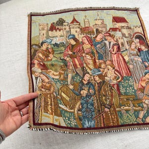 May include: A tapestry depicting a medieval scene with figures in period clothing, set against a backdrop of castles and buildings. The tapestry features a burgundy border and a fringed edge, showcasing a detailed woven design.