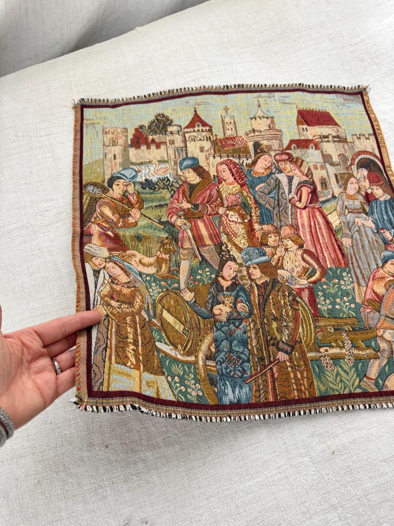 May include: A tapestry depicting a medieval scene with figures in period clothing, set against a backdrop of castles and buildings. The tapestry features a burgundy border and a fringed edge, showcasing a detailed woven design.