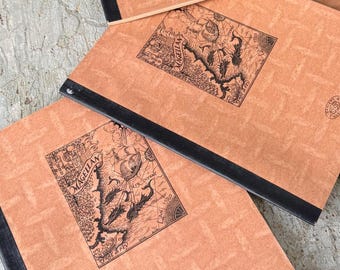 May include: Several notebooks with a vintage map design. The covers are a light brown color with a black binding. The map design is black and features the word "Magellan" and the year "1520". The notebooks are stacked on top of each other.