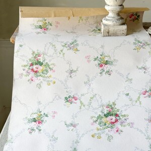 Könnte beinhalten: Eine Rolle Vintage-Tapete mit floralem Muster. Das Design zeigt Gruppen von rosa, roten, gelben und grünen Blumen mit hellblauen Ranken auf cremefarbenem Hintergrund. Die Tapete ist teilweise abgerollt.